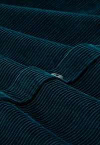 Teal corduroy stof met een opvallende ribbelstructuur, gevouwen met een zichtbare knop, die een golvende uitstraling creëert in zacht, pluizig materiaal.