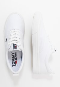 Białe sneakersy z płótna z gumową podeszwą, sznurowanym designem i markową wkładką z napisem "TOMMY JEANS" w czerwonych i niebieskich akcentach.