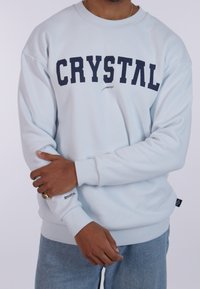 Felpa blu cielo con la scritta "CRYSTAL" in grassetto lettere navy, polsini a costine e una texture morbida in misto cotone. Vestibilità casual con dettagli del marchio.
