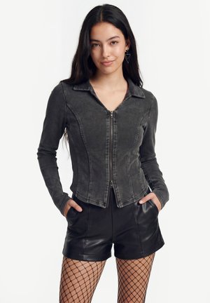 Fiatal nő hosszú sötét hajjal, visel egy testhezálló sötétszürke cipzáras dzsekit, fekete bőr rövidnadrágot és hálós harisnyát, kezét zsebre téve.