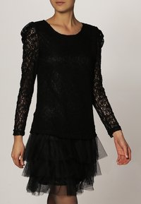 Top de encaje negro de manga larga con cuerpo texturizado, escote redondo y falda de tul con volantes en capas, creando una apariencia similar a un vestido.