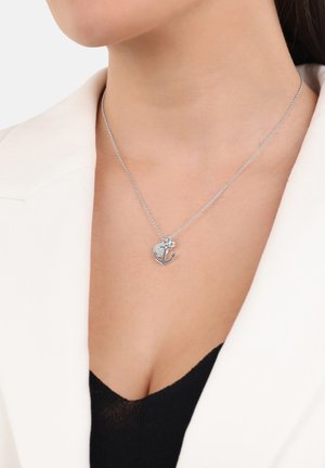 Collana in argento con un pendente che combina un cuore texturizzato e un'ancora, indossata con una camicia nera e un blazer bianco.