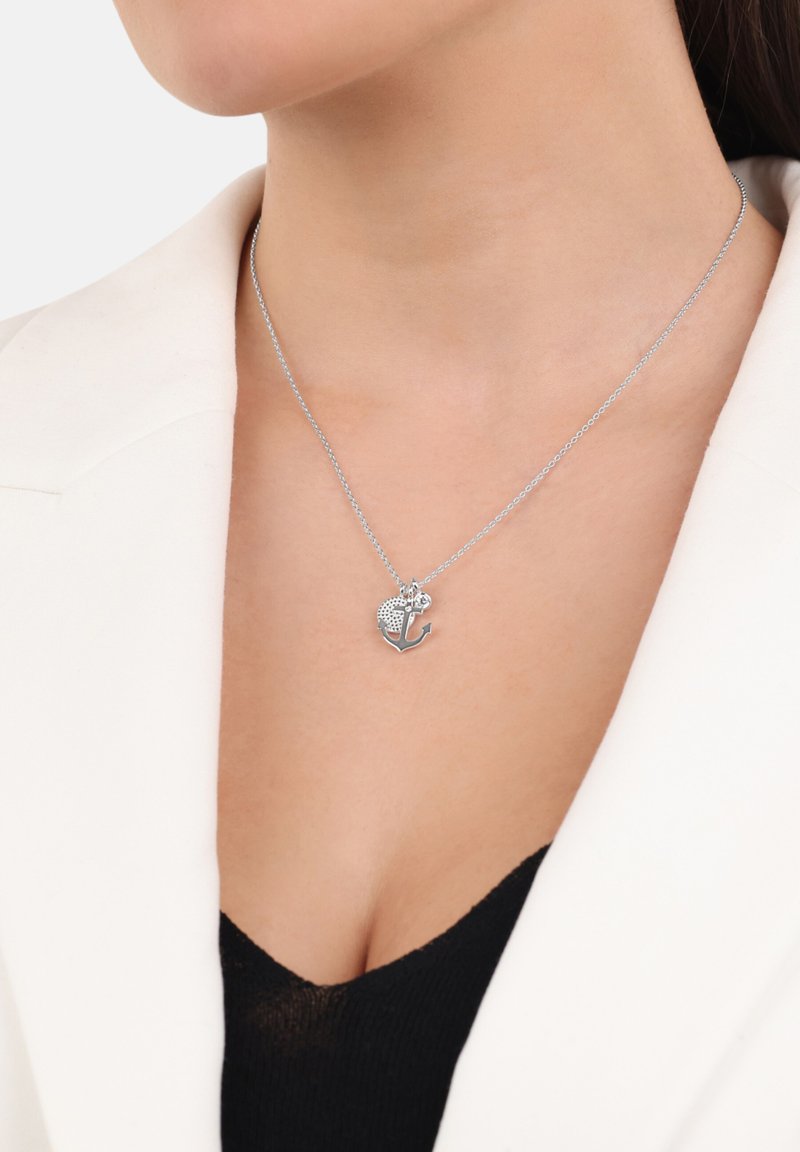 Collana in argento con un pendente che combina un cuore texturizzato e un'ancora, indossata con una camicia nera e un blazer bianco.