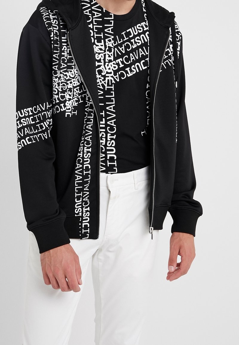 Homme portant un sweat à capuche zippé noir et un T-shirt noir avec le texte blanc répété "JUST CAVALLI", associé à un pantalon blanc sur un fond uni.