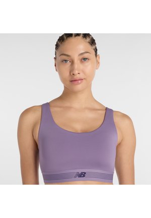 Jeune femme aux cheveux tressés portant un soutien-gorge de sport violet clair avec un logo New Balance foncé sur fond blanc.