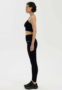 Czarny sportowy biustonosz i legginsy z wysokim stanem przedstawione w profilu. Legginsy z logo, oba wykonane z elastycznego materiału. Czarne obuwie sportowe.