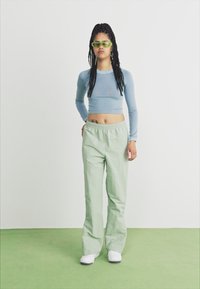 Camisa de manga larga azul clara, pantalones deportivos verdes y zapatillas blancas. El modelo lleva gafas de sol verdes. Tejido suave y corte relajado.