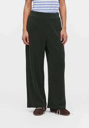 Personne portant un pantalon large vert foncé, des chaussures marron avec des lacets blancs et une chemise rayée bleue et blanche, debout sur un fond blanc.