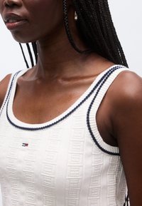 Bílý pletený nátělník s texturovaným vzorem, s námořnicky modrými akcenty podél výstřihu a ramínek. Malé logo na přední straně.