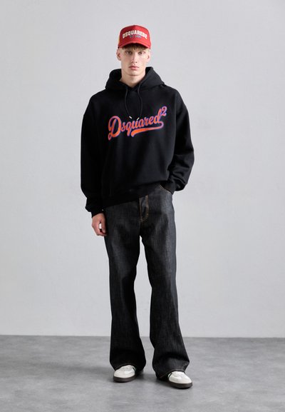 Svart huvtröja med "Dsquared²" i blå och röd text, kombinerad med mörk denimjeans och en röd keps med varumärket.