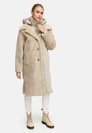Manteau en fausse fourrure beige avec un col à revers, boutons croisés, poches latérales, superposé sur une doudoune beige et un pantalon blanc, associé à des bottes beiges.