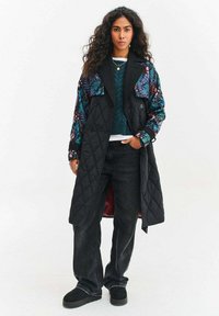 Prešívaný čierny trenčkot s farebnými kvetinovými rukávmi, ktorý má veľké vrecká a červenú vnútornú podšívku, nosený cez zelený sveter a čierne džínsy.