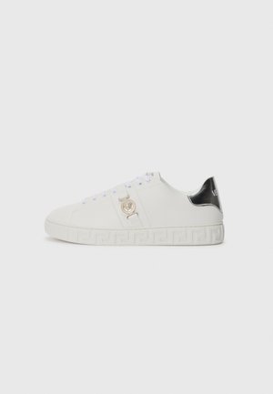 Zapatilla blanca de cuero con puntera redondeada, acento de logotipo dorado y plantilla de talón negra. Patrón decorativo a lo largo de la suela blanca.