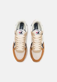 Beige and tan sneakers with a mesh and suede upper, navy accents, white laces, and a branded tongue label. Features a textured rubber sole.
