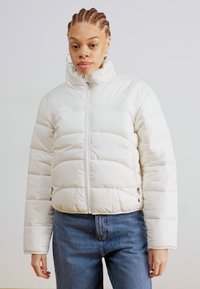 Hvit pufferjakke med høy krage, glidelås og polstret design. Har sidelommer og en kort modell. Bært med blå jeans.