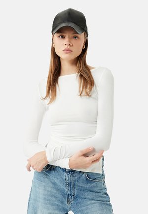 Jonge vrouw met lang bruin haar, een zwarte leren pet, een wit shirt met lange mouwen, blauwe spijkerbroek en gouden oorringen, staand met gekruiste armen.