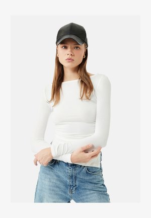 Jonge vrouw met lang bruin haar, een zwarte leren pet, een wit shirt met lange mouwen, blauwe spijkerbroek en gouden oorringen, staand met gekruiste armen.