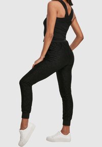 Femme portant un pantalon de jogging noir texturé avec les chevilles retroussées, un haut sans manches noir et des baskets blanches, debout avec une jambe levée.