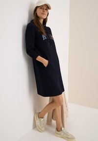 Mörkblå huvtröja med ficka, med kontrasterande bokstavstryck; kombinerad med ljusa sneakers och en beige cardigan. Mjuk textur.