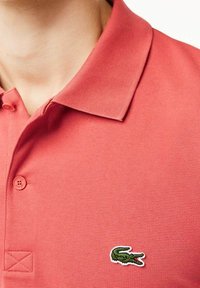 Röd polo med en strukturerad tyg, som har en klassisk krage, två knappar och en grön broderad krokodil-logotyp på bröstet.