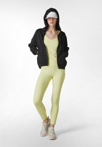 Zwarte rits hoodie, lichtgele tanktop en leggings. De outfit heeft een gladde textuur. Lichte sneakers met groene accenten maken de look compleet.