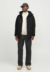 Schwarze Kapuzenjacke mit zwei Reißverschlusstaschen, cremiger Sweatshirt mit hohem Kragen, dunkle Hose mit Cargotaschen und beigefarbene Stiefel.