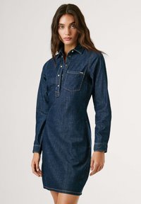 Tume sinine teksakleid, millel on pikad varrukad, nööpidega krae ja rinnatasku. Omab nähtavat õmblust ja kitsast lõike.