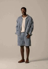 Stribet blå og hvid kortærmet jakke med matchende shorts, struktureret beige sweater og brune sandaler; afslappet pasform og knapdetaljer.