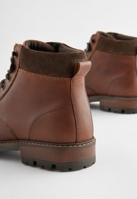 Hnedé kožené členkové topánky so semišovým golierom, guľatým tvarom, šnurovaním vpredu a robustnou, textúrovanou podrážkou. Dve ťahacie pútka na zadnej strane.