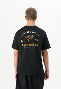 Mann med kort hår som har på seg en svart Ripcurl Surf Co t-skjorte med kenguru og Australia tekst på ryggen, står mot en hvit bakgrunn.