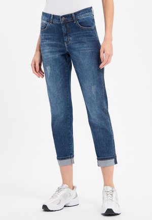 Femme portant un jean bleu retroussé avec une légère usure et des baskets blanches, debout devant un fond blanc uni.