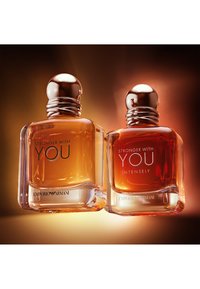 Due flaconi di profumo Emporio Armani etichettati "Stronger With You" e "Stronger With You Intensely" con tappi metallici su uno sfondo caldo.