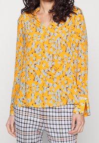 Kvinde iført en langærmet gul bluse med blå blomsterprint og rutede bukser i hvide, sorte og orange nuancer.