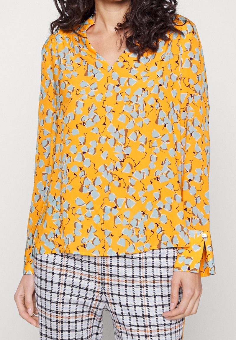 Kvinde iført en langærmet gul bluse med blå blomsterprint og rutede bukser i hvide, sorte og orange nuancer.