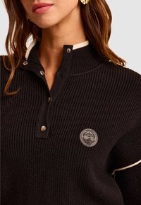 Czarny sweter o prążkowanej strukturze z wysokim kołnierzem, z kremowym akcentem, napami i okrągłą naszywką na lewej piersi.