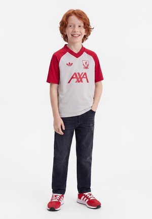 Rødhåret dreng smiler, iført en hvid og rød Liverpool fodboldtrøje, mørke jeans og røde sneakers, står med den ene hånd i lommen.