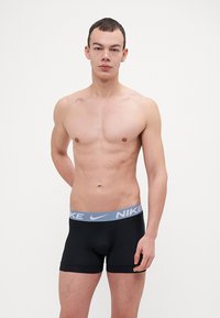 Crne bokserice s laganim sivim pojasom s Nike logom. Glatka tkanina, uski dizajn i dužina srednje dužine. Model stoji s neutralnim izrazom lica.