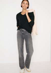 Schwarzer strukturiertes V-Ausschnitt-Pullover, graue gerade Jeans und helle Sneakers, mit einer cremefarbenen Jacke über einem Arm drapiert.