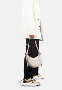 Borsa a mano beige chiaro con forma curva, texture in pelle liscia e manico corto, tenuta da una persona che indossa un blazer bianco e pantaloni a righe.
