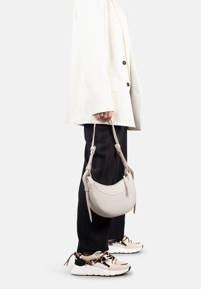 Borsa a mano beige chiaro con forma curva, texture in pelle liscia e manico corto, tenuta da una persona che indossa un blazer bianco e pantaloni a righe.