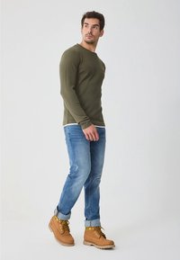 Lang groen olijfkleurig hemd met lange mouwen, blauwe spijkerbroek en beige laarzen. Het hemd heeft een eenvoudig ontwerp; de laarzen hebben gele veters en een robuuste zool.