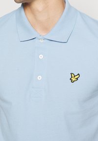Polo shirt azzurro chiaro con tessuto strutturato, tre bottoni bianchi e un piccolo logo di un uccellino giallo ricamato sul petto.