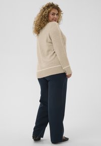Pull beige avec une finition texturée et des poignets côtelés, associé à un pantalon large bleu marine et des chaussures plates noires, vu de dos.