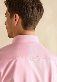 Ružová košeľa s golierom, hladkou textúrou a štítkom "JOULES" na zadnej časti výstrihu. Košeľa má klasický strih a dizajn.