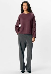 Maroonowy sweatshirt z dekoracyjnymi wzorzystymi akcentami na rękawach, połączony z szarymi dopasowanymi spodniami i czarnymi butami na szpilkach.