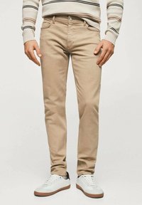Hombre que lleva pantalones ajustados beige, zapatillas blancas con suelas marrones, y un suéter beige con rayas negras y marrones en las mangas.
