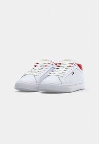 Balsvos baltos sportbačiai su baltais raišteliais, raudona vidine pamušalu ir Tommy Hilfiger logotipu ant liežuvėlio ir šono, eksponuojami ant baltos fono.