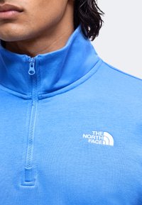 Gros plan d’une personne portant une veste bleu quart de zip avec le logo The North Face sur la poitrine, fermeture éclair tirée jusqu’au col.