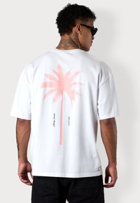 Weißes Baumwoll-T-Shirt mit einem großen, pinken Palmenbaum-Design auf der Rückseite. Das Design enthält Text in der Nähe der Basis der Grafik.