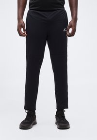 Άτομο που φοράει μαύρο αθλητικό παντελόνι Nike Pro και μαύρα αθλητικά παπούτσια, στέκεται μπροστά σε απλό λευκό φόντο.